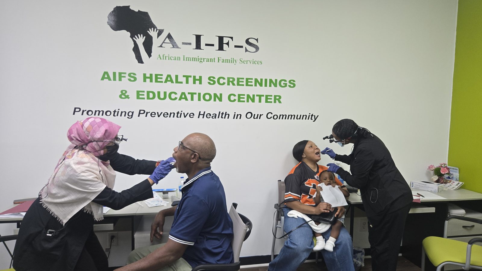 Two screening activities take place at tables, with healthcare workers attending to seated adults in a room with AIFS signage.